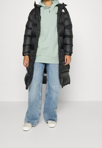 Cappotto piumino nero con cappuccio, caratterizzato da una texture lucida e ampie sezioni trapuntate. Indossato sopra un hoodie verde chiaro e jeans a zampa d'elefante blu.