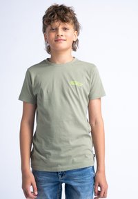 Olijfgroen katoenen T-shirt met ronde hals, korte mouwen en een kleine neonafbeelding op de borst. Gecombineerd met blauwe denim jeans.