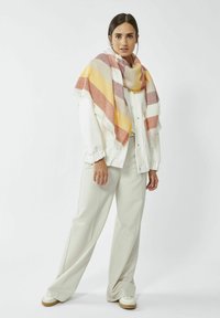 Grote sjaal met een ruitpatroon in aardse tonen van oranje, geel en crème, gedrapeerd over een witte blouse en grijze broek. Met franjes aan de randen.