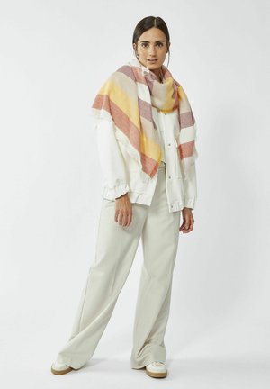 Grote sjaal met een ruitpatroon in aardse tonen van oranje, geel en crème, gedrapeerd over een witte blouse en grijze broek. Met franjes aan de randen.