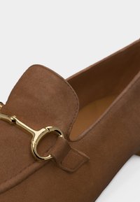 Mocassin en daim marron avec détail en métal doré en forme de mors de cheval sur le devant, doté d'un bout arrondi et d'un talon bas.