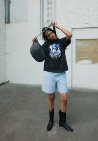 Black oversized t-shirt with graphic print, light blue checkered shorts, gray patterned handbag, and black cowboy boots. Neutral background.