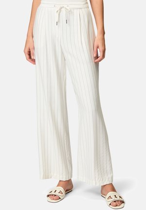 Pantalon blanc à jambes larges avec de fines rayures verticales noires, taille à cordon, porté avec des sandales plates crème à bouts ouverts sur une personne debout.