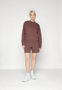 Pruun puuvillane pusa langevate õlgadega, sobitatud pruuni lühikeste pükstega. Jalal valged spordijalatsid ja heledad sokid.