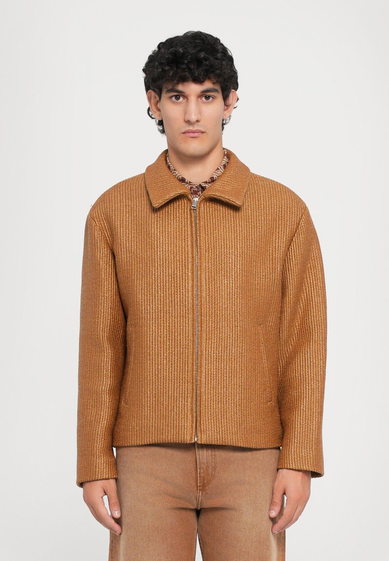 Chaqueta marrón de canalé con cierre zip y cuello, de mangas largas y diseño ajustado, combinada con pantalones marrones a juego. Detalles de textura y costura notables.