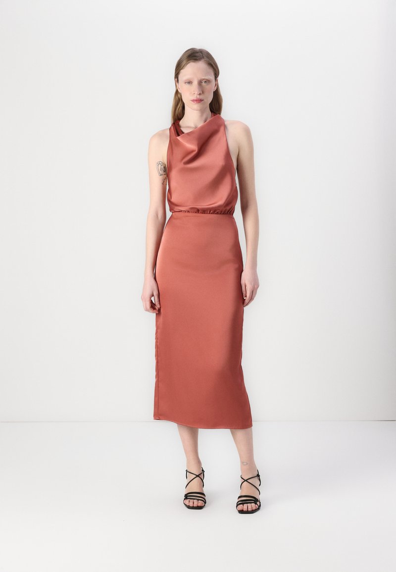 Abito in satin color ruggine con scollo a cowl, vita aderente e silhouette dritta, abbinato a sandali neri con tacco alto e fasce.