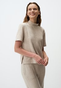 Top beige a maniche corte con colletto alto, caratterizzato da una texture morbida e spacchi laterali. Abbinato a pantaloni beige coordinati.