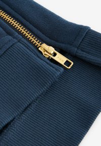 Zippad huva i mörkblått tyg. Har en guldfärgad metalldragkedja och ribbad textur längs ärmsluten och fållen.