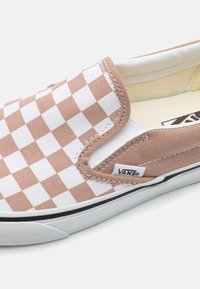 Zapato slip-on de lona de cuadros marrón y blanco, con suela negra, costuras blancas y una etiqueta con el logo de Vans en el lateral.