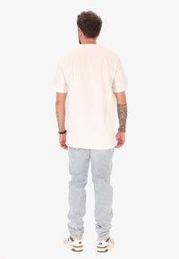 Homme avec des tatouages sur les deux bras portant un t-shirt blanc uni, un jean bleu clair et des baskets blanches, se tenant de dos sur un fond blanc.