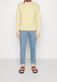 Sudadera amarilla clara, vaqueros azules y sandalias marrones. La sudadera tiene un cuello redondo y puños acanalados. Los vaqueros están remangados en el dobladillo.