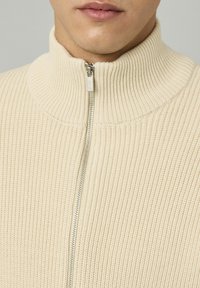 Homme portant un pull en tricot côtelé couleur crème avec une fermeture éclair argentée partiellement zippée au cou.