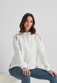 Woman with shoulder-length brown hair wearing a white zip-up sweatshirt with red stripes and blue leggings, seated against a plain background.