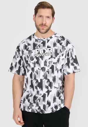 Mężczyzna o jasnobrązowych włosach i brodzie, ubrany w czarno-białą koszulkę z wzorem miasta i napisem „FOR SNWVGEAR”, stoi z jedną ręką w kieszeni.