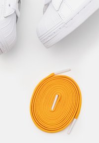 Vita sneakers med texturerat läderovandel och gummisula, kombinerat med en gul, rund skosnöre med plastaglets i båda ändar.