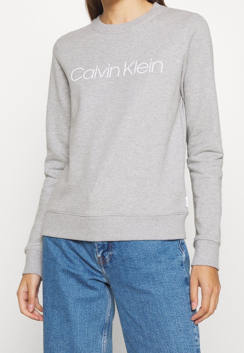 Personne portant un sweat-shirt gris clair Calvin Klein avec un logo texte blanc et un jean bleu, debout devant un fond uni.