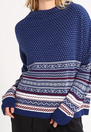 Sininen neulepaita, jossa on geometrisia kuvioita valkoisena ja punaisena, leveä pyöreä kaula-aukko, pitkät hihat sekä ribbaatut hihansuut ja helma.