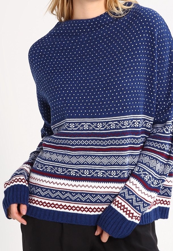 Niebieski sweter z dzianiny z geometrycznymi wzorami w bieli i czerwieni, szerokim okrągłym dekoltem, długimi rękawami oraz ściągaczami na mankietach i u dołu.