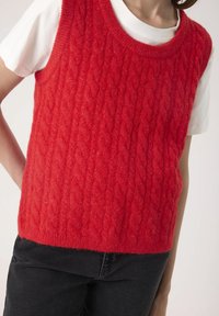 Red cable-knit sleeveless sweater worn over a white short-sleeve shirt with black pants, showing torso and partial arms.