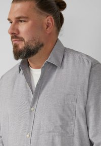 Camicia a maniche lunghe grigia con motivo, colletto, taschino sul petto e tessuto testurizzato, indossata sopra una maglietta bianca.