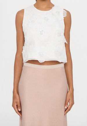 Mouwloze witte crop top met grote bloemenborduursel gecombineerd met een hoog opgesneden lichtroze rok, die de taille blootlegt.