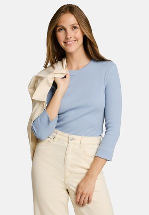 Jeune femme aux longs cheveux châtain clair portant une chemise bleu clair à manches longues et un pantalon crème, tenant une veste crème sur son épaule.