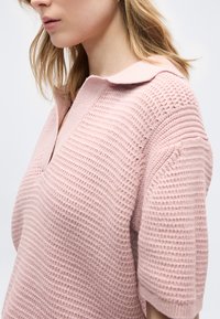 Kvinde iført en lyserød strikket kortærmet sweater med en opklappet krave, vist fra siden i brysthøjde.