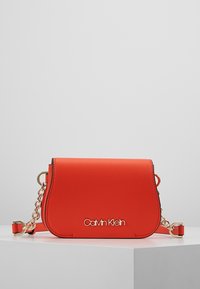 Sac bandoulière en simili cuir rouge avec garnitures dorées. Il présente un rabat incurvé, un accent en chaîne et un logo embossé sur le devant.