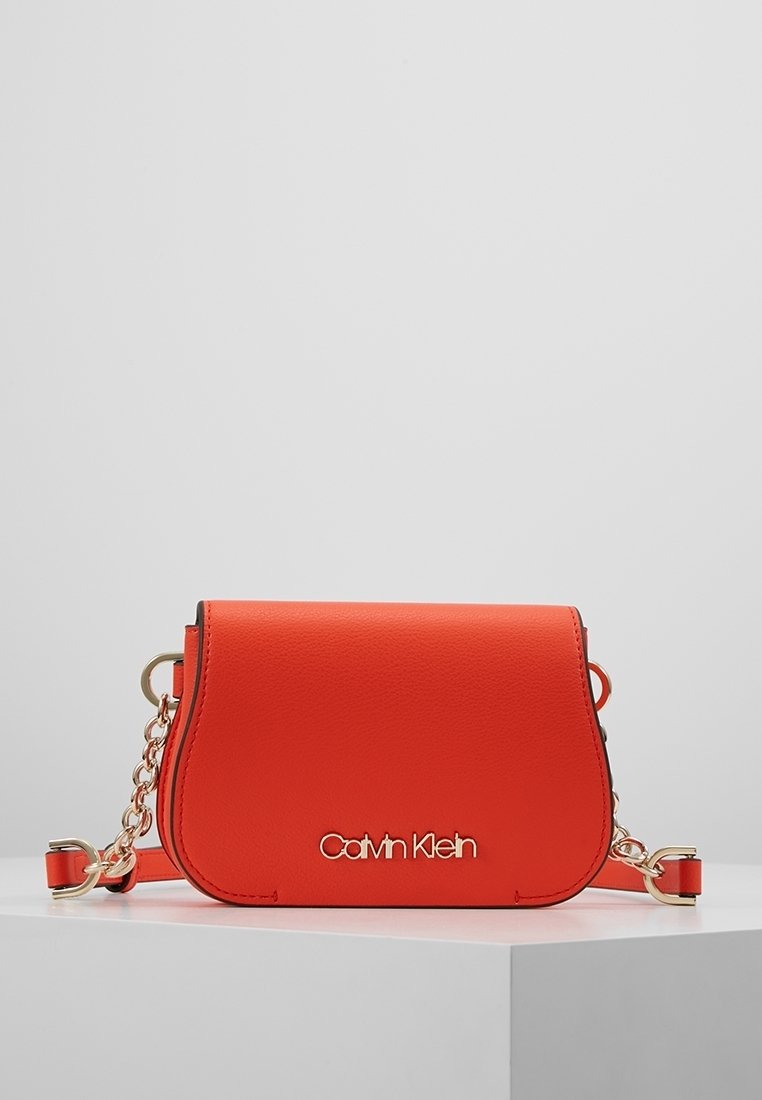 Sac bandoulière en simili cuir rouge avec garnitures dorées. Il présente un rabat incurvé, un accent en chaîne et un logo embossé sur le devant.