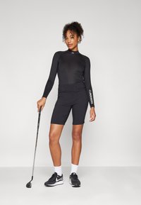 Svart langermet topp og tettsittende svarte shorts, begge laget av elastisk stoff. Modellen holder en golfklubb, iført svarte joggesko og hvite sokker.