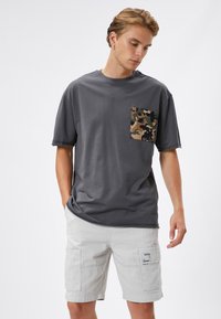 Camiseta gris de manga corta con un bolsillo de camuflaje. La tela es suave; presenta un corte relajado y un ligero desgaste en los bordes de las mangas.