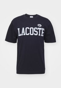 Tummansininen puuvillapaita, jossa on rohkea valkoinen "LACOSTE" -teksti ja pieni Lacoste-logo vasemmalla puolella. Lyhyet hihat ja klassinen istuvuus.