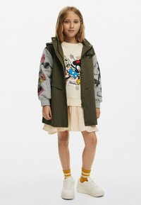 Olivgrön parkas med grå mönstrade ärmar, lager på lager över en krämfärgad grafisk tröja och ljusrosa kjol, matchad med vita sneakers och randiga strumpor.