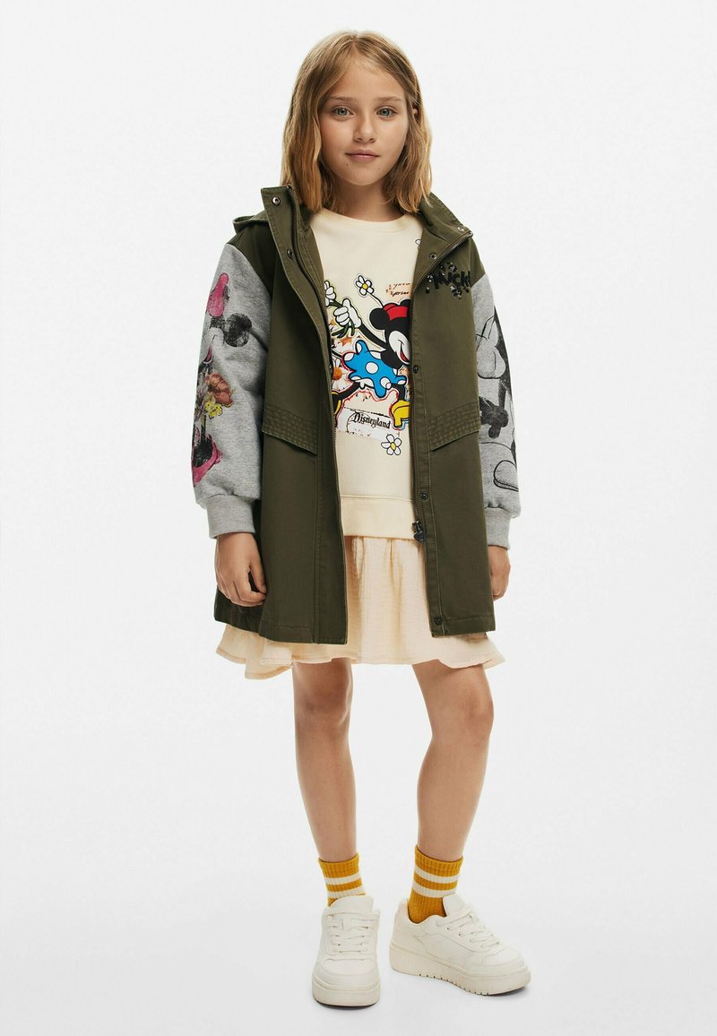 Olivgrön parkas med grå mönstrade ärmar, lager på lager över en krämfärgad grafisk tröja och ljusrosa kjol, matchad med vita sneakers och randiga strumpor.