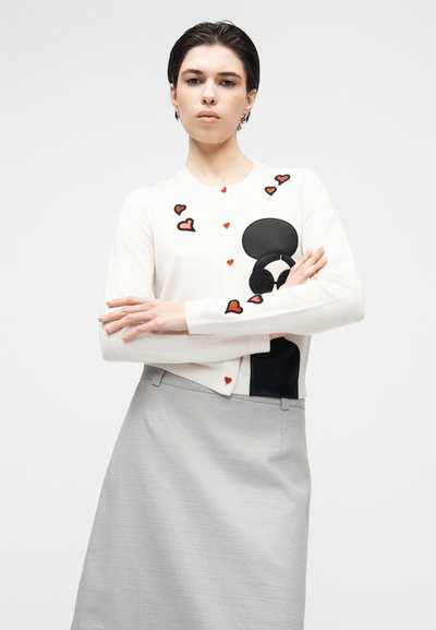 Fehér kardigán piros szívmintákkal és fekete díszítésekkel, gombos záródással és bordázott mandzsettákkal, szürke szoknyával párosítva.