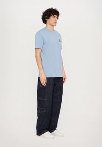Tricou din bumbac de culoare albastru deschis, cu mâneci scurte și un buzunar pe piept, purtat cu pantaloni cargo din denim închis, având buzunare laterale și cusături de contrast.