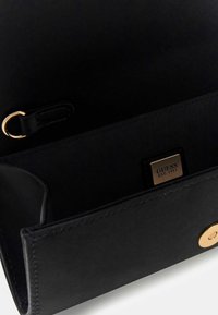 Interno di una borsa in pelle nera con texture liscia; presenta un accento in metallo dorato e una targhetta con il marchio "GUESS EST. 1981."