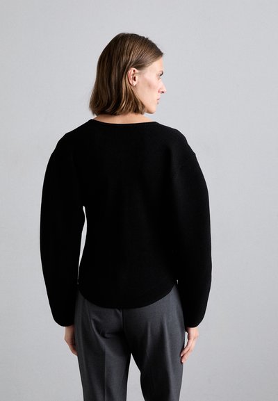 Black ribbed knit sweater with wide, long sleeves and a rounded neckline, paired with gray tailored trousers. Textured fabric visible.