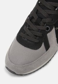 Chaussure de sport grise combinant mesh et daim, bout arrondi, accents noirs, et lacets plats. Motif texturé sur les côtés.