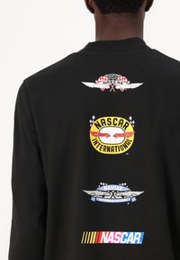 Svart langermet skjorte med flere fargerike NASCAR-logoer på baksiden, inkludert vinge-design og striper i forskjellige farger.