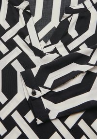 Zwart-wit gebloemde blouse met geometrische vormen, een V-halslijn en een knoopmanchet. Gemaakt van gladde stof met een zachte textuur.