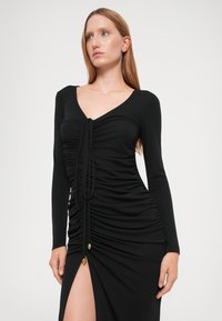 Black long-sleeve dress with a V-neck, featuring gathered texture and a front tie. Includes a thigh-high slit and gold accent details.