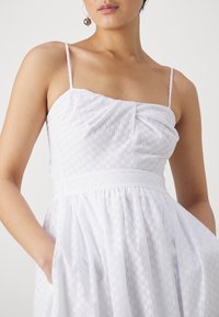 Γυναίκα που φοράει ένα αμάνικο λευκό φόρεμα με λεπτές τιράντες και διακριτό υφαντό σχέδιο, με τα χέρια στις τσέπες, σε απλή φόντο.