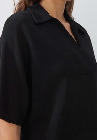 Černá košile s krátkým rukávem a límečkem, vyrobená z texturované látky a vybavená náprsní kapsou. Design je volnější, s uvolněnou siluetou.