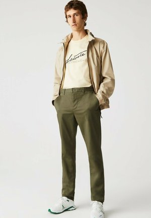 Veste légère beige avec une fermeture éclair, t-shirt crème avec un texte noir, pantalon vert olive et chaussures blanches avec des accents verts.