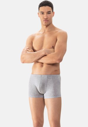 Muskelöser Mann steht mit verschränkten Armen da, trägt enganliegende graue Boxershorts vor hellem einfarbigem Hintergrund.