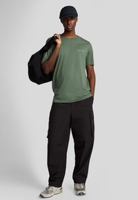 Grøn kortærmet skjorte med logo, sorte cargo-bukser og en sort kasket. Holder en sort duffelbag. Grå baggrund. Afslappet outfit.