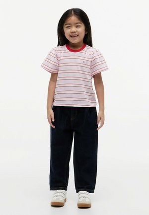 Młoda dziewczyna uśmiechająca się, ubrana w białą koszulkę z cienkimi czerwonymi paskami, ciemne szerokie spodnie i białe trampki, stojąca na jednolitym tle.