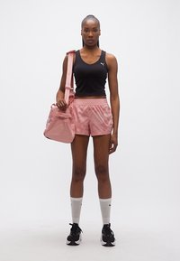 Γυναίκα με μαύρο αμάνικο τοπ και ροζ σορτς που κρατά ροζ τσάντα που ταιριάζει, φορά λευκές κάλτσες και μαύρα αθλητικά παπούτσια, στέκεται μπροστά σε λευκό φόντο.