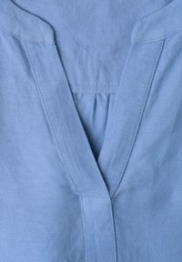 Chemise en lin bleu clair avec col en V et coutures doubles visibles le long du col et de l'encolure.